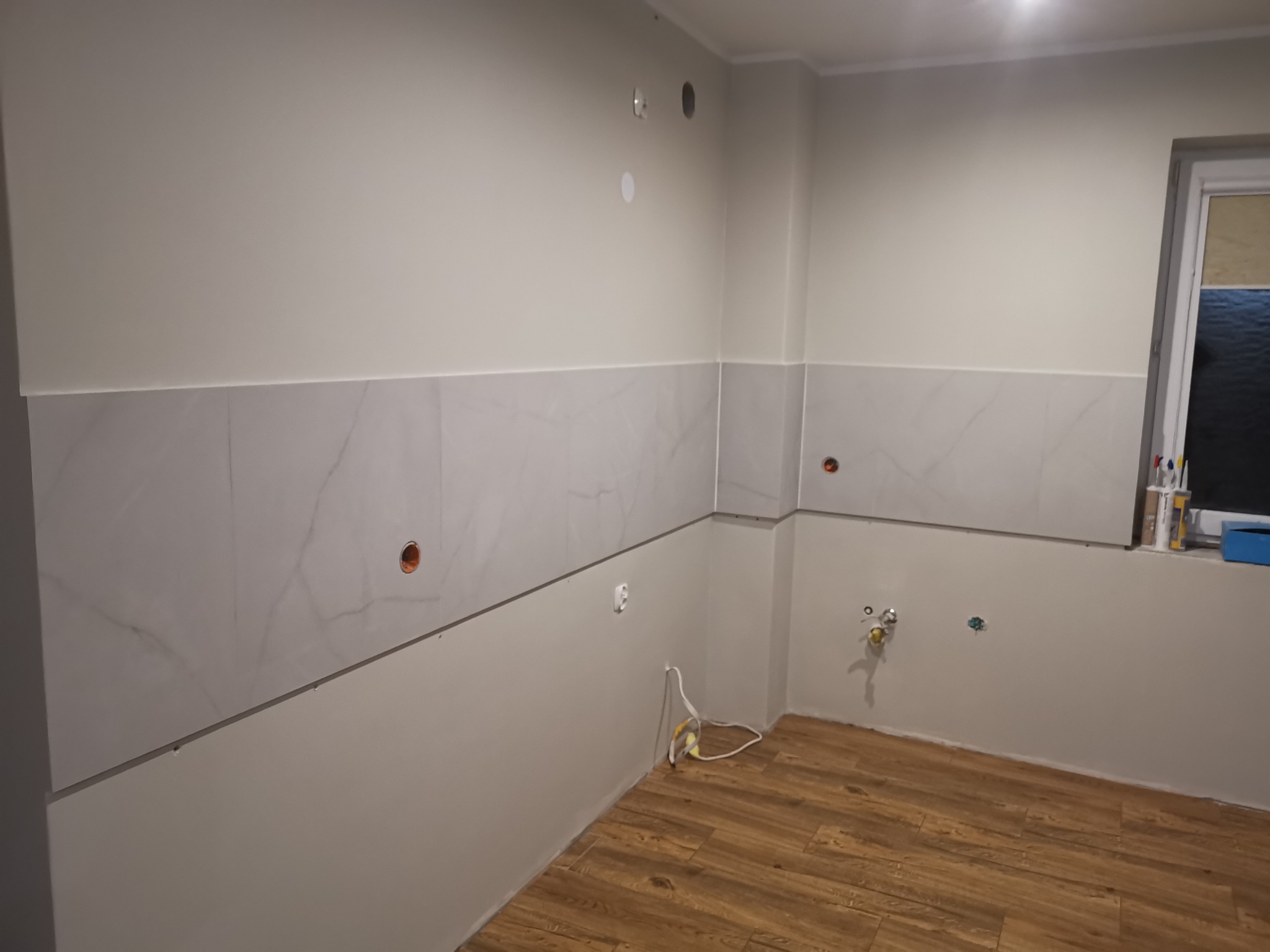 Fragment wnętrza w trakcie prac. Częściowo położone płytki imitujące marmur na ścianie, widoczne instalacje elektryczne i hydrauliczne, podłoga wyłożona panelami.