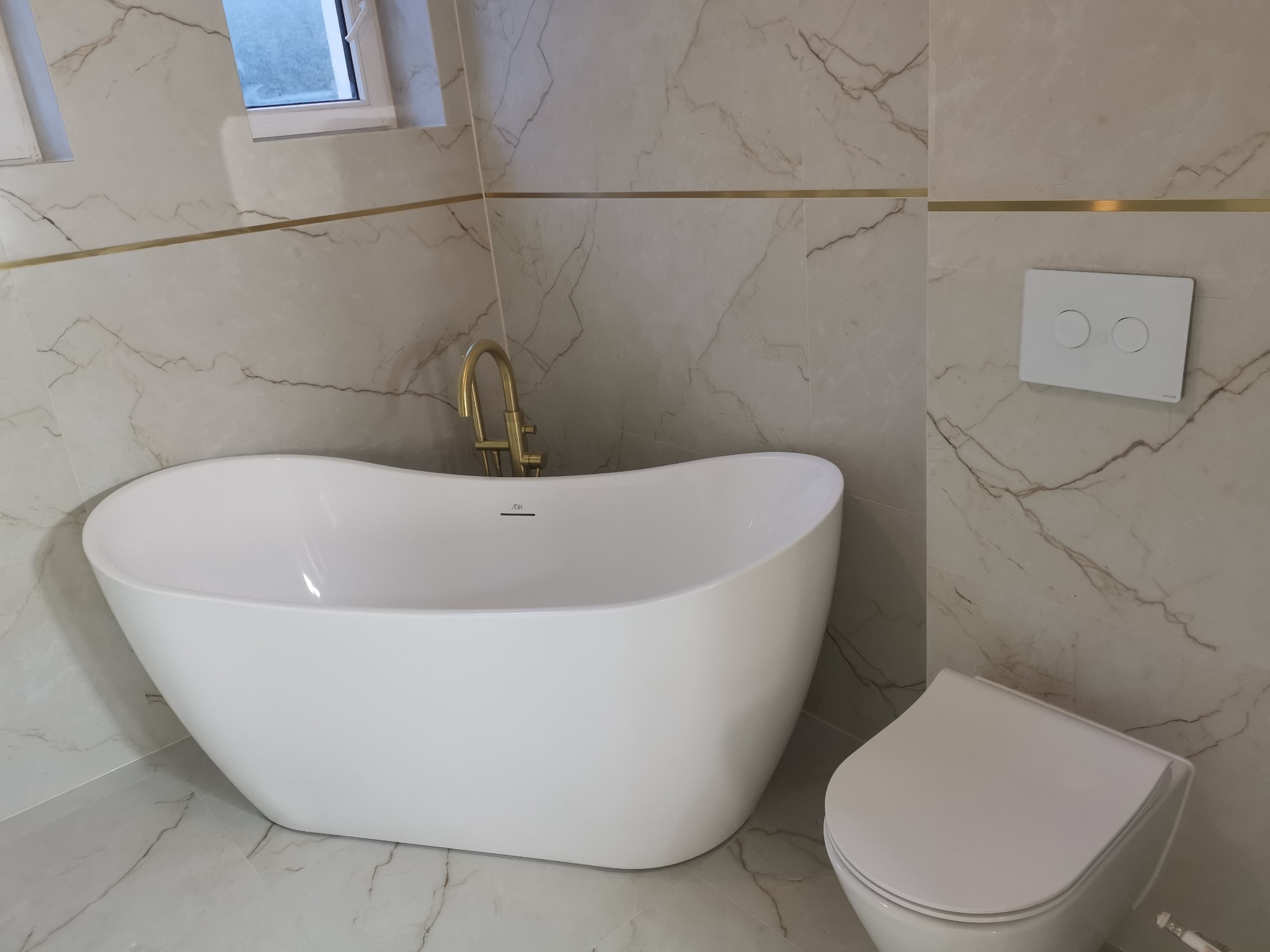 Biała, wolnostojąca wanna i toaleta w nowoczesnej łazience z marmurowymi płytkami i złotymi akcentami. Minimalistyczny design, jasne wnętrze.