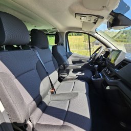 GEAR SHIFTERS EUROPE SPÓŁKA Z OGRANICZONĄ ODPOWIEDZIALNOŚCIĄ - Wnętrze busa z szarymi fotelami, widok z perspektywy pasażera. Na zewnątrz zielony krajobraz z domem i ogrodzeniem. Jasne, słoneczne oświetlenie.