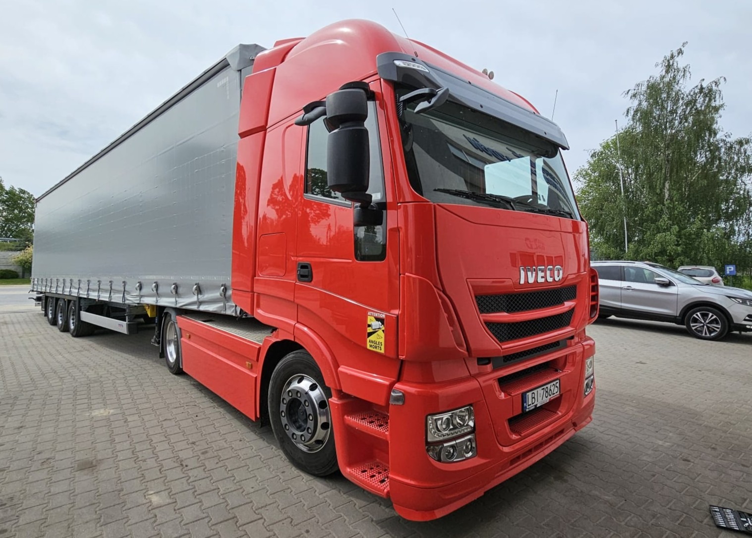 Czerwona ciężarówka Iveco z naczepą na parkingu, widoczny fragment tablicy rejestracyjnej. W tle drzewo i zaparkowane samochody osobowe.