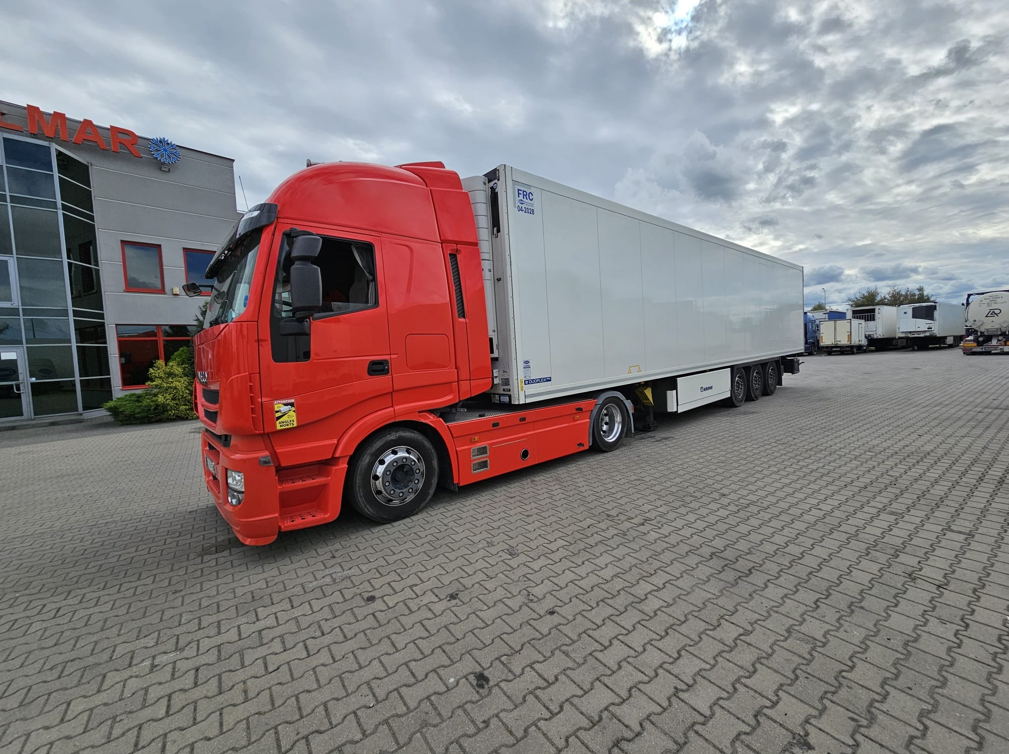 Czerwona ciężarówka chłodnia marki Iveco, zaparkowana na placu z kostki brukowej, z budynkiem firmy w tle i pochmurnym niebem.