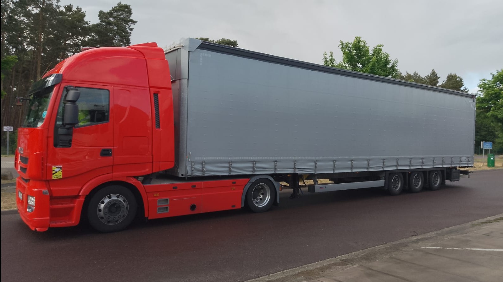 Czerwona ciężarówka z szarą naczepą na asfalcie, widok z boku, drzewa w tle. Tabliczka ostrzegawcza 'Angles Morts' na kabinie.