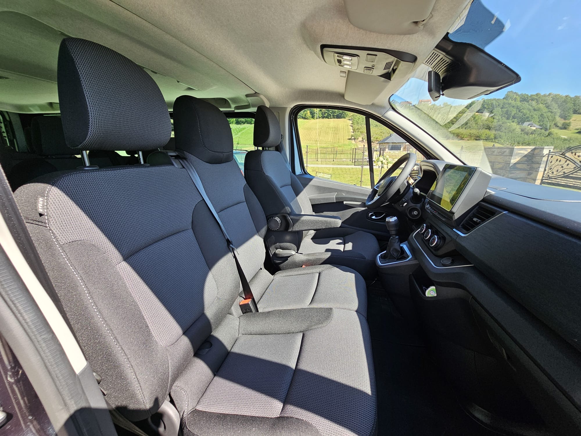 Wnętrze busa z szarymi fotelami, widok z perspektywy pasażera. Na zewnątrz zielony krajobraz z domem i ogrodzeniem. Jasne, słoneczne oświetlenie.