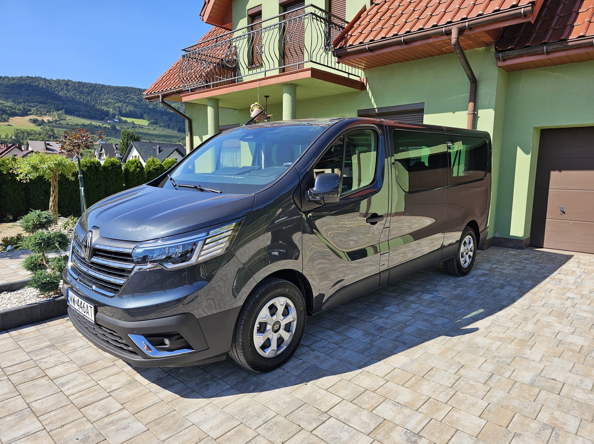 Szary bus Renault Trafic na tle domu z zieloną elewacją i brązowym dachem, zaparkowany na kostce brukowej w słoneczny dzień. Widok z przodu i boku.