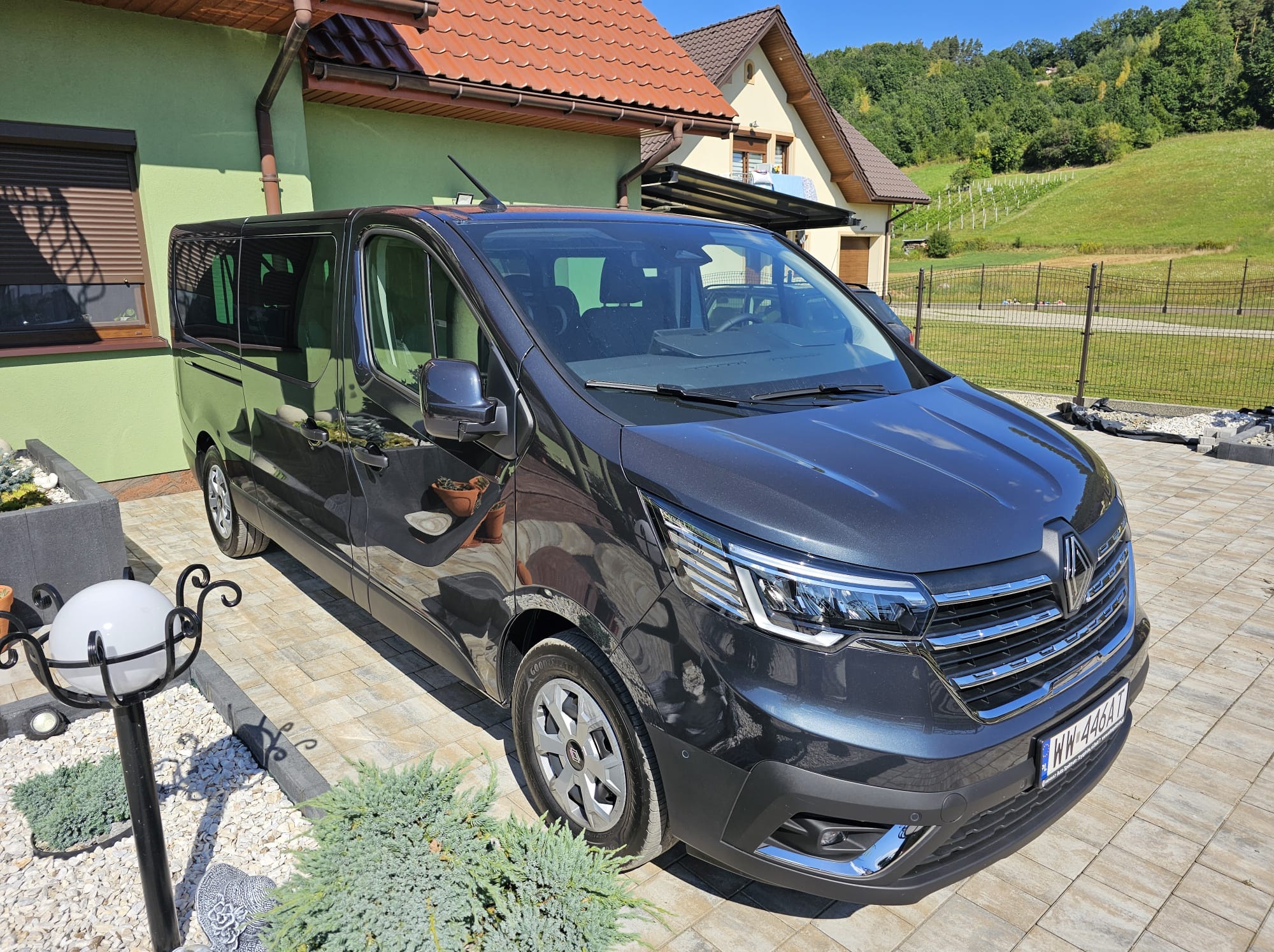 Szary Renault Trafic na tle domu z czerwoną dachówką i zielonego trawnika. Auto zaparkowane na kostce brukowej, widoczna tablica rejestracyjna.