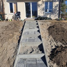 Patryk Żołnowski - Schody z kostki brukowej prowadzące do domu, otoczone ziemią. Widoczne okna i balkon domu w tle. Słoneczny dzień, świeżo wykonane prace ziemne i brukarskie.