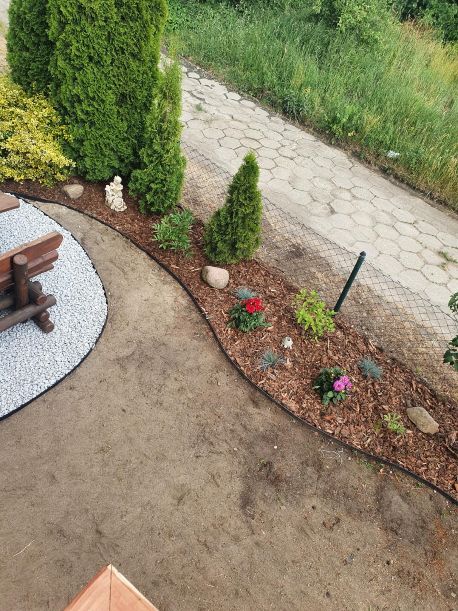 Aranżacja ogrodu z rabatą kwiatową, obrzeżem i ścieżką z białego żwiru. W tle widoczny chodnik z kostki i fragment ogrodzenia z siatki.
