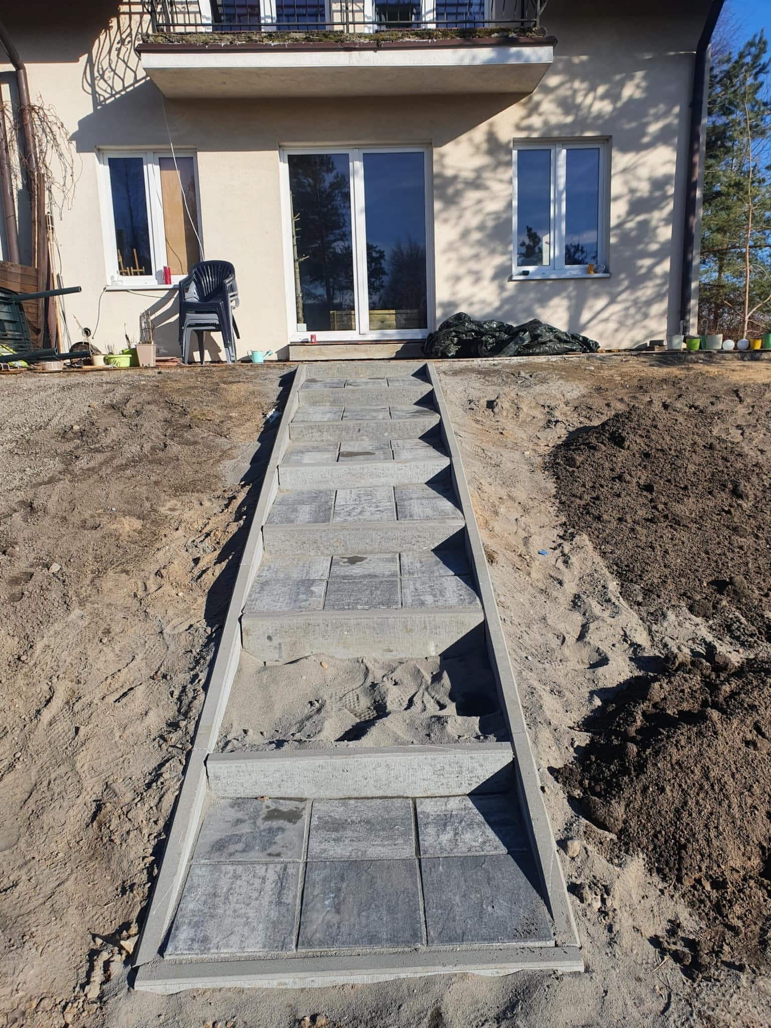 Schody z kostki brukowej prowadzące do domu, otoczone ziemią. Widoczne okna i balkon domu w tle. Słoneczny dzień, świeżo wykonane prace ziemne i brukarskie.