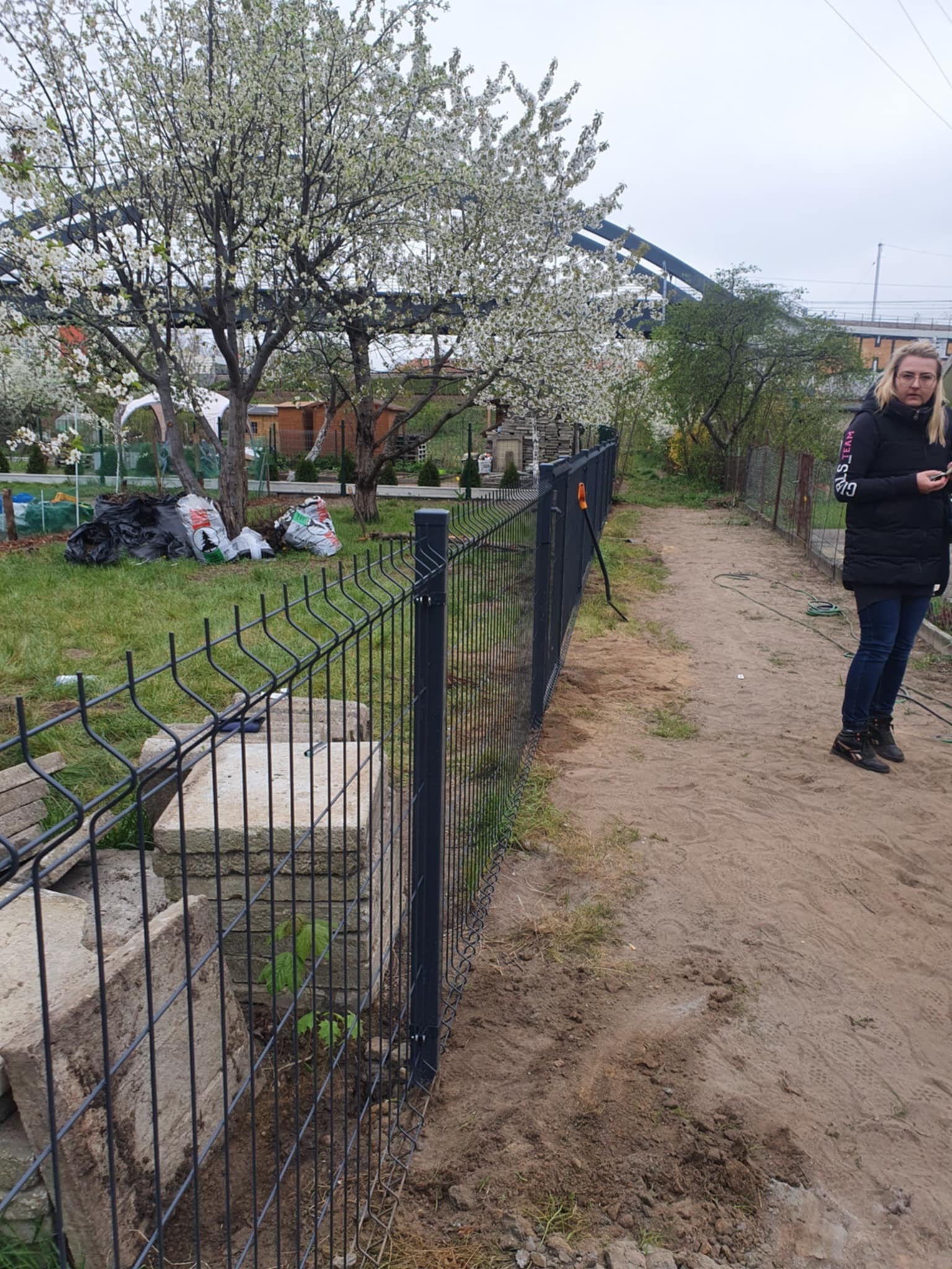Częściowo zamontowane, grafitowe ogrodzenie panelowe na piaszczystym podłożu, z widocznymi słupkami i betonowymi elementami. W tle kwitnące drzewo i kobieta.