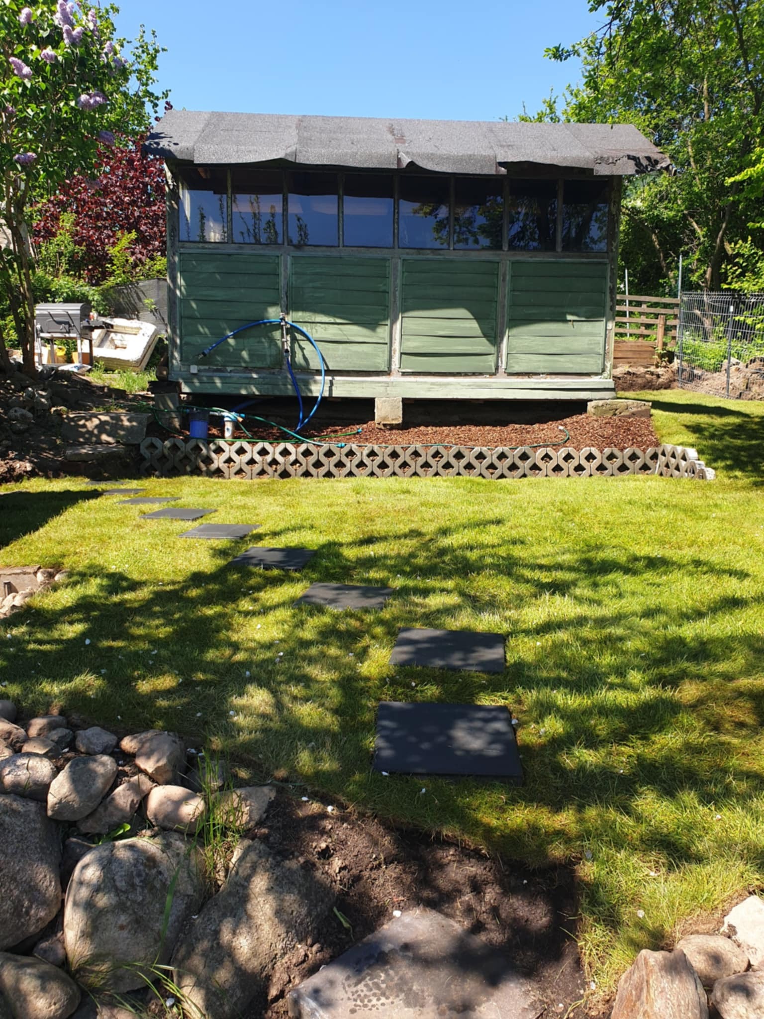 Zielony domek narzędziowy z oknami, otoczony trawnikiem i ścieżką z płyt. Dekoracyjne obrzeże z betonowych pustaków. W tle drzewa i krzewy.