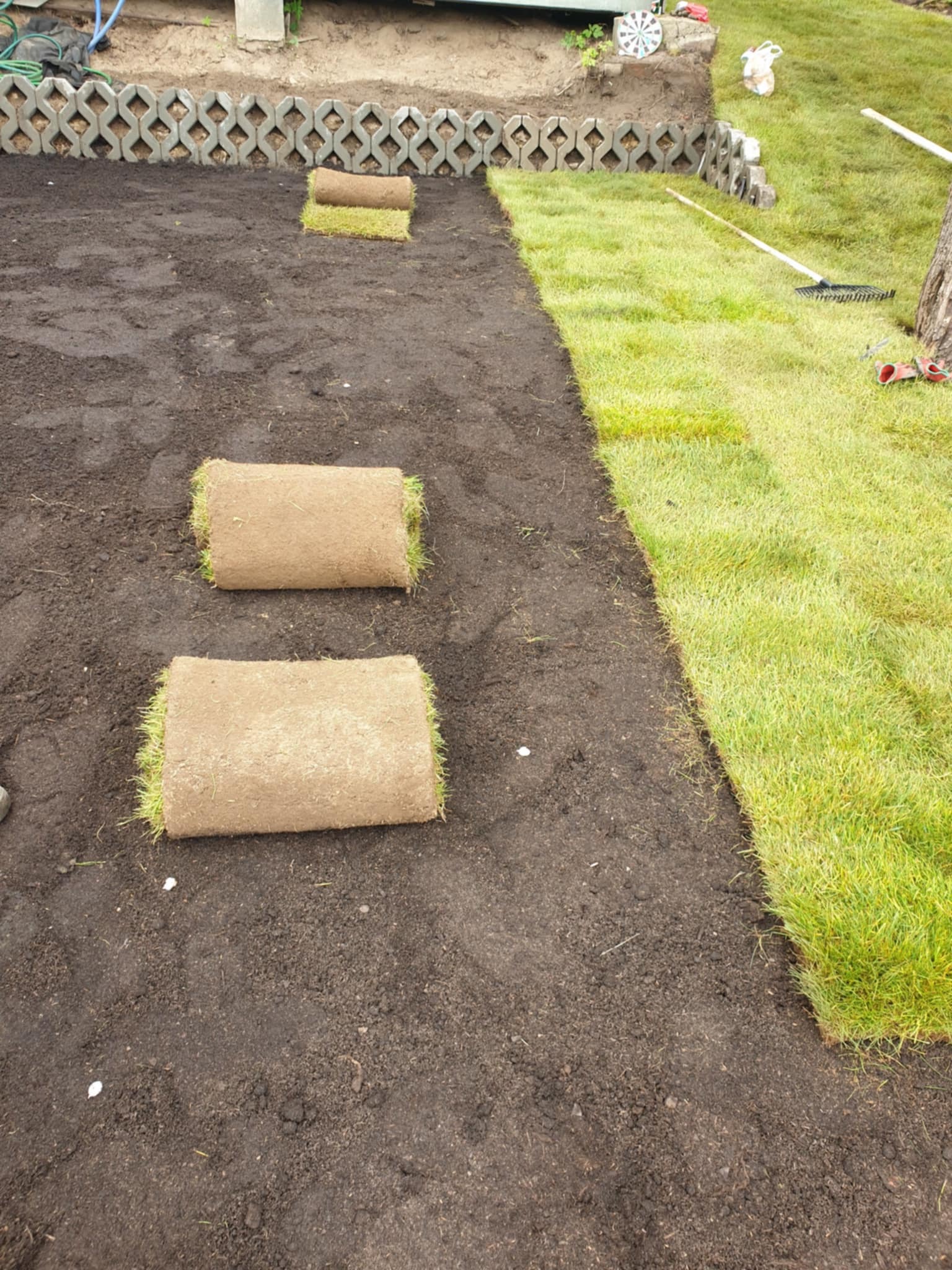 Rozkładanie rolek trawy na przygotowanym podłożu, widoczny fragment ułożonego trawnika i dekoracyjny murek z betonowych elementów w tle.