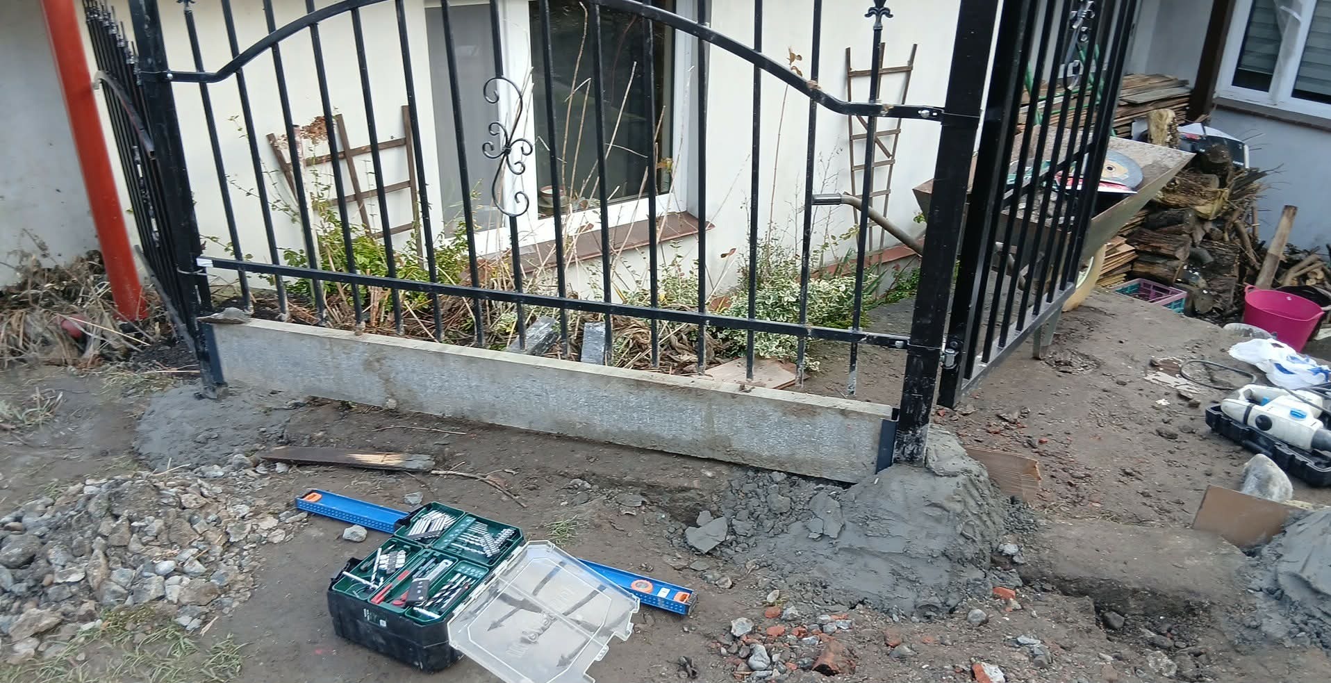 Fragment montażu czarnego, kutego ogrodzenia z widocznymi narzędziami i betonową podstawą w trakcie prac ziemnych. W tle biały budynek i składowane drewno.