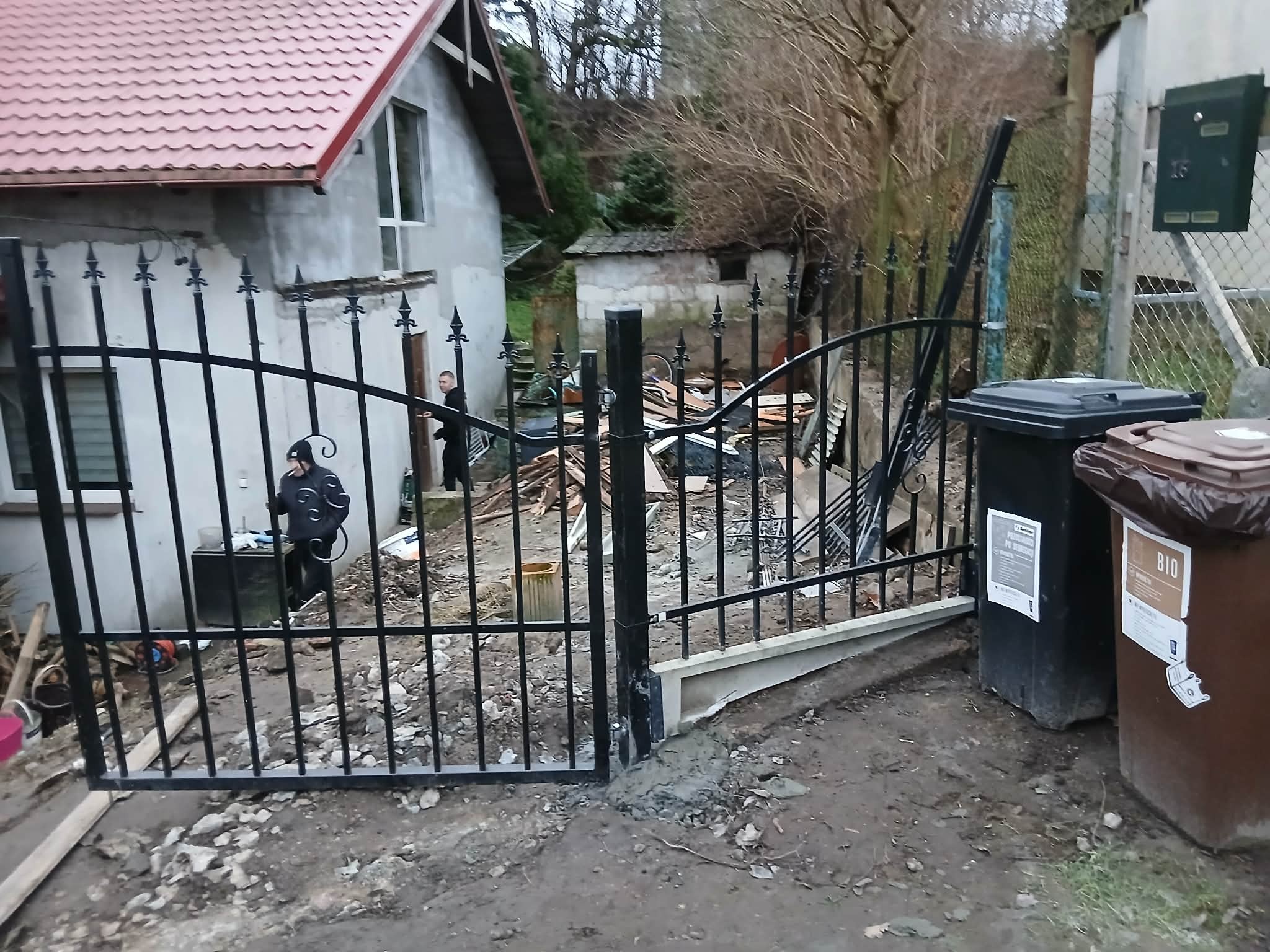 Montaż czarnego, metalowego ogrodzenia z ozdobnymi grotami. Widoczne dwa kosze na śmieci i fragment domu w tle. Prace montażowe w toku, teren wokół rozkopany.