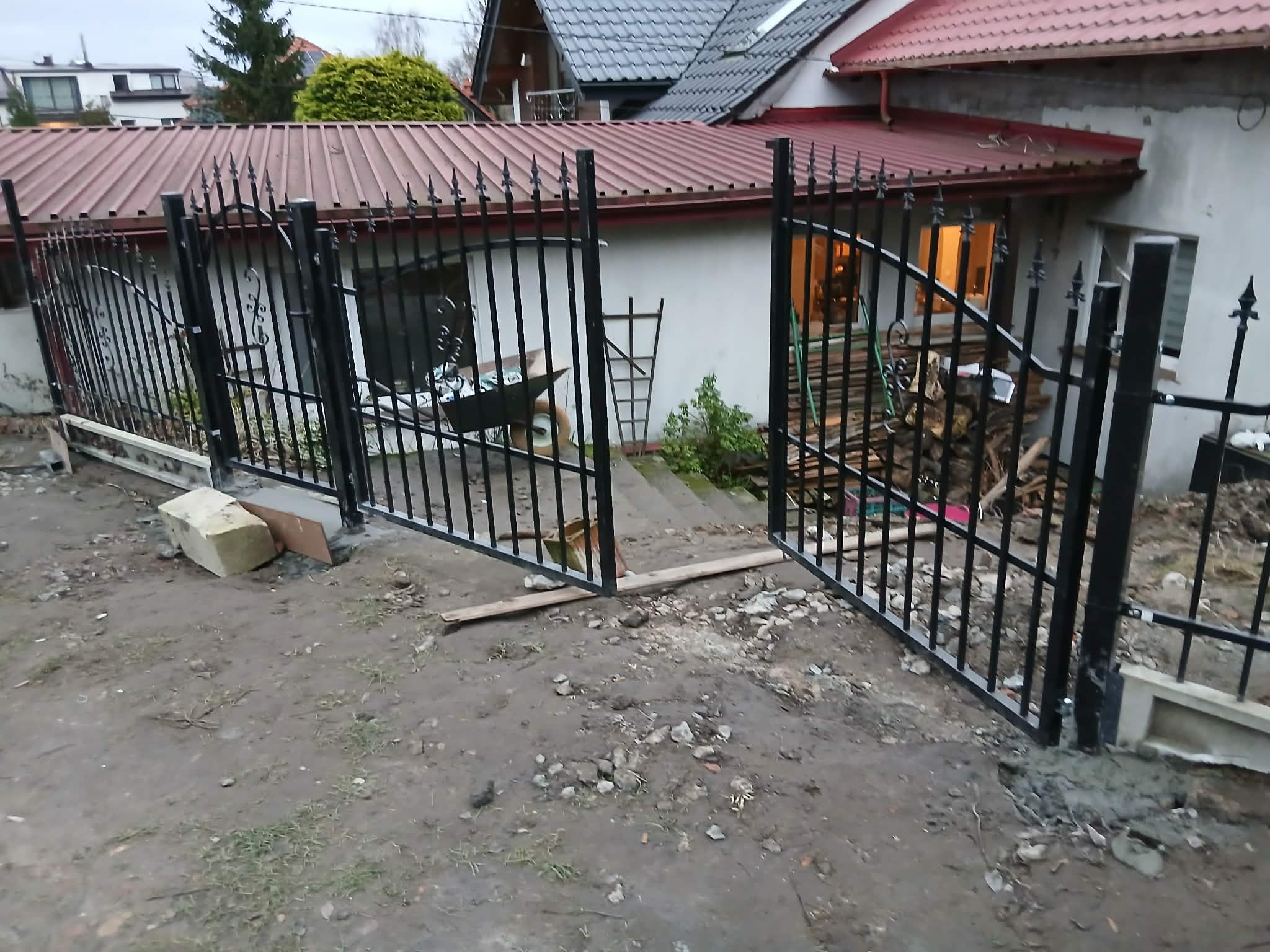 Częściowo zamontowane, czarne, metalowe ogrodzenie z otwartą bramą na posesji. Widoczne elementy montażowe i teren po budowie. W tle dom z czerwoną dachówką.