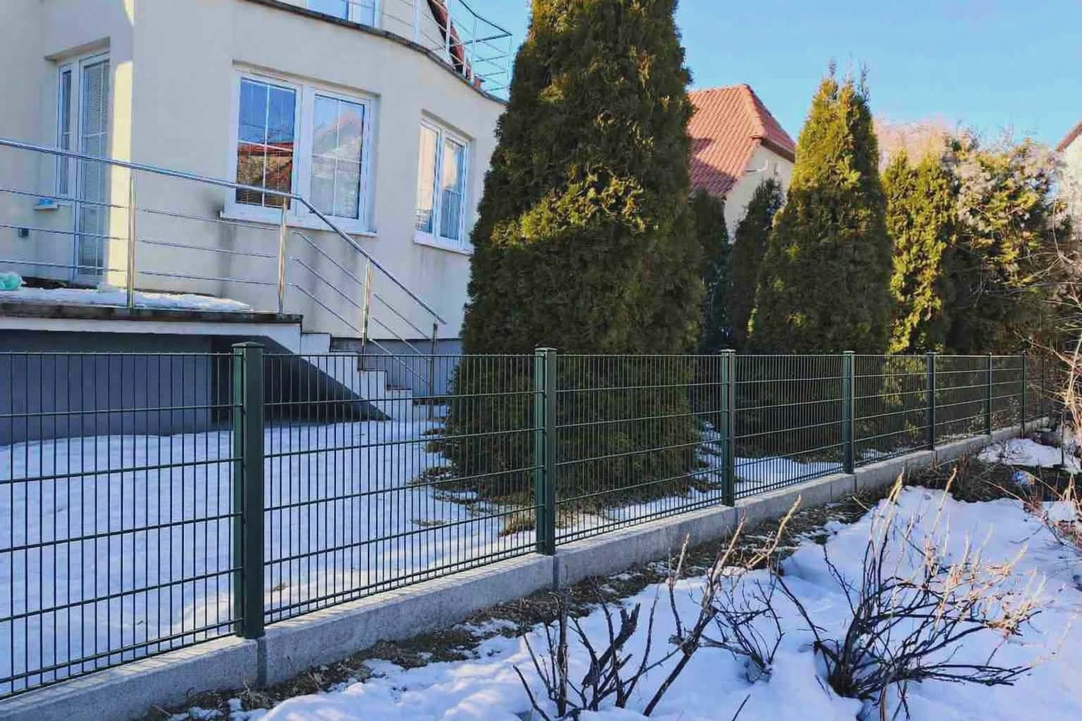 Nowoczesne ogrodzenie panelowe w kolorze zielonym, zamontowane na betonowej podmurówce, z widocznym budynkiem w tle i resztkami śniegu na trawniku.