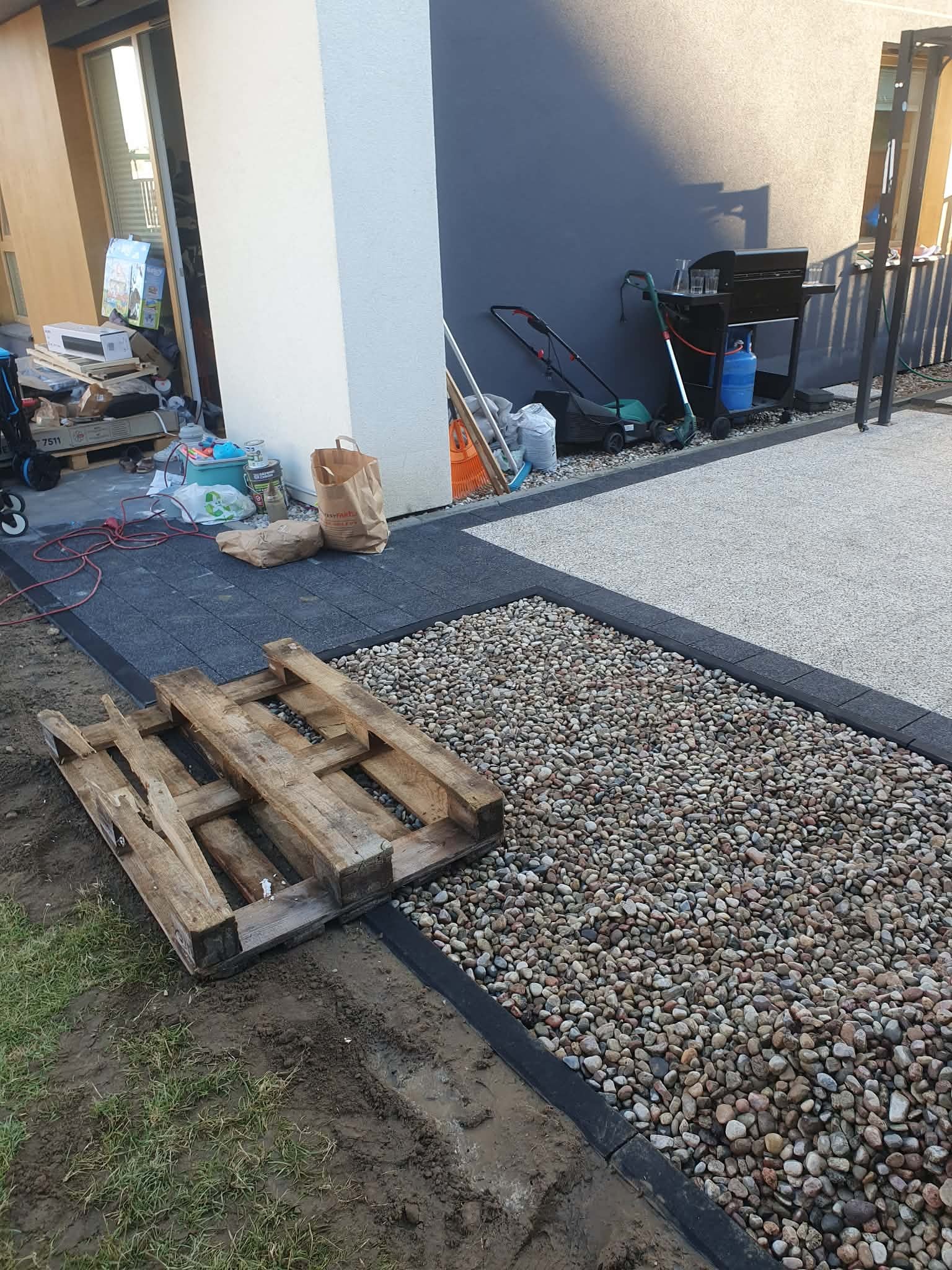 Fragment ogrodu w trakcie budowy: ułożone kamienie, kostka brukowa, paleta drewniana. W tle dom z grillem i kosiarką. Widoczny etap prac ziemnych i układania nawierzchni.