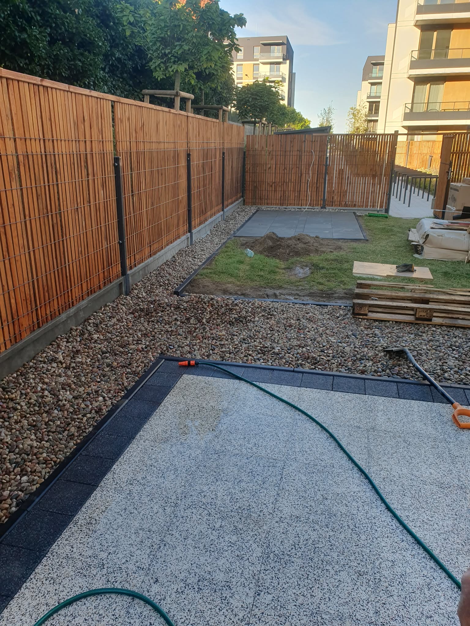 Nowoczesne ogrodzenie panelowe z drewnianym wypełnieniem, oddzielające posesję. Widoczny fragment tarasu z kamienną nawierzchnią i zielony wąż ogrodowy.