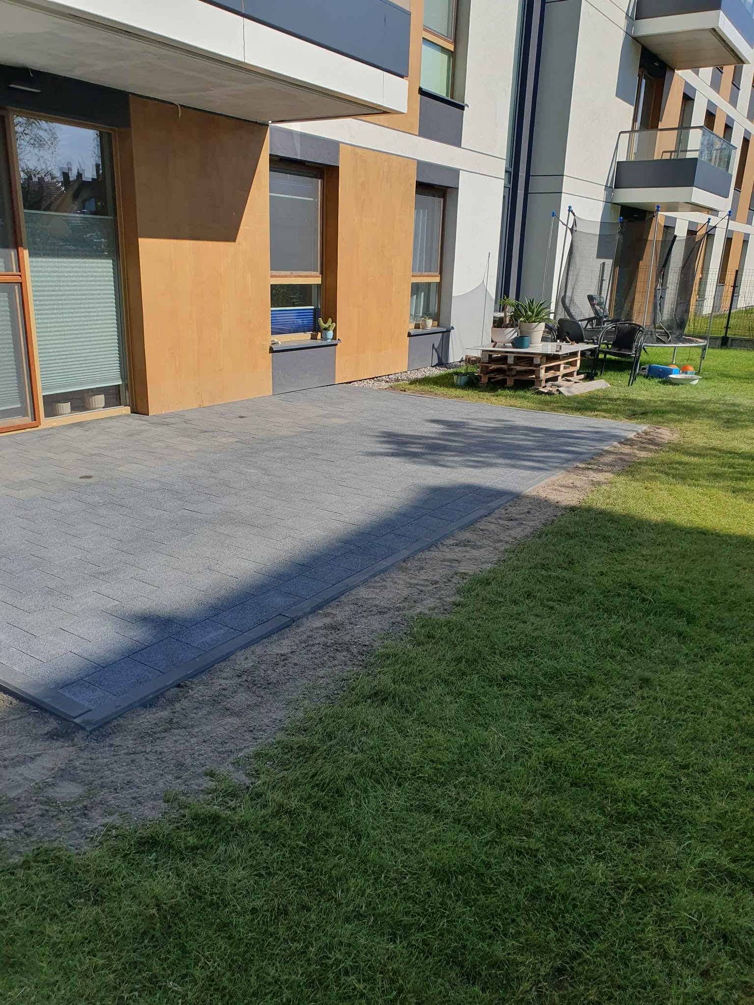 Ułożona kostka brukowa na posesji przy budynku wielorodzinnym, widok na trawnik i elementy wyposażenia ogrodu, takie jak trampolina i stolik z palet.