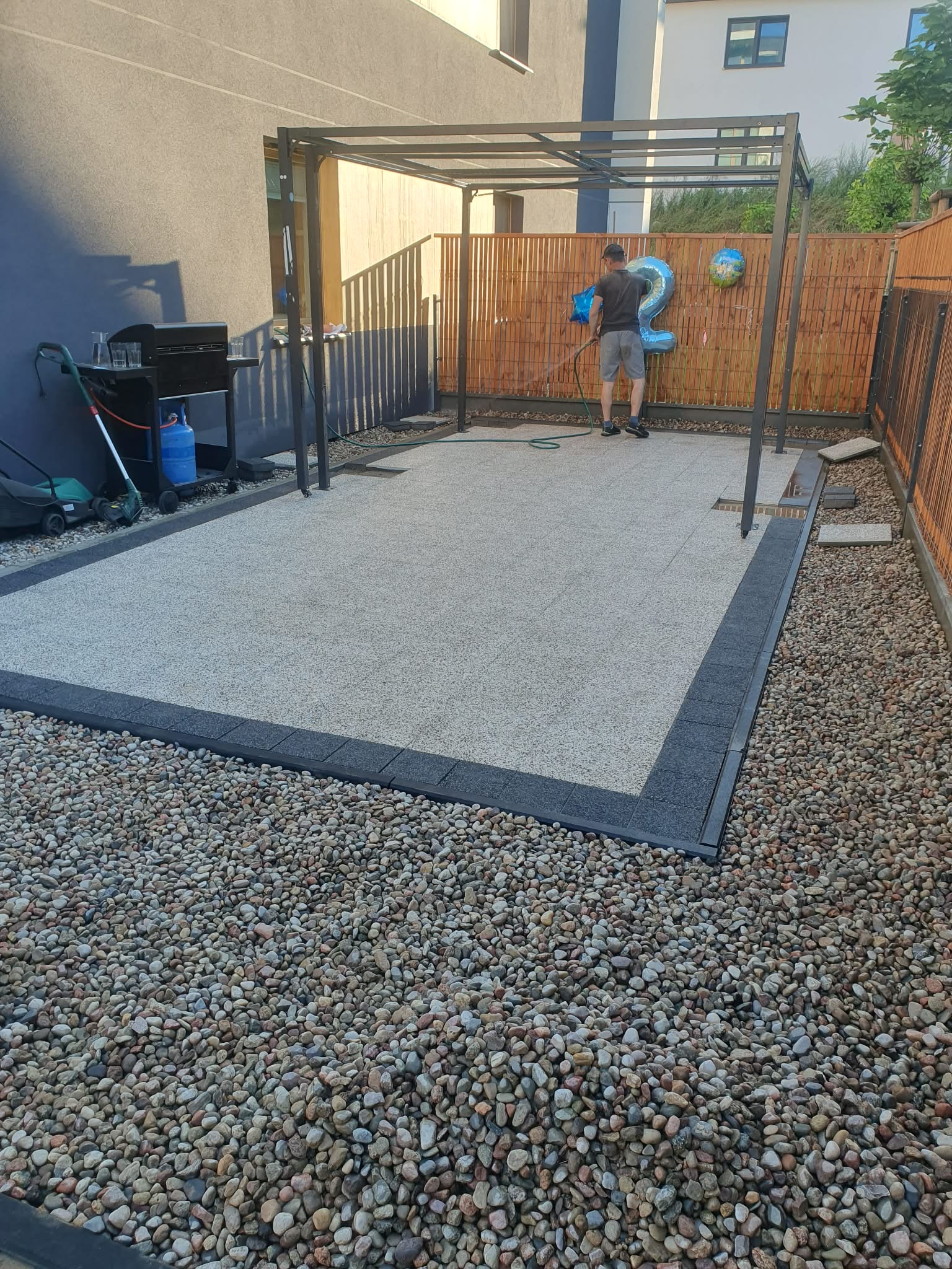 Wykończony taras z kamienną nawierzchnią i obrzeżami, z pergolą i fragmentem ogrodzenia. Mężczyzna podlewa teren w tle, widoczne dekoracje urodzinowe.