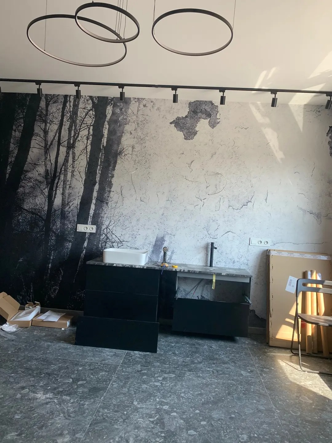 Częściowo zmontowana czarna szafka łazienkowa z umywalką na tle ściany z motywem lasu i surowym wykończeniem. Widoczne elementy oświetlenia.