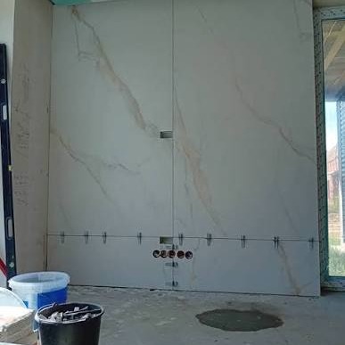 Ściana w trakcie wykańczania z dużymi płytami imitującymi marmur, widoczne otwory na gniazdka i poziomica. Etap prac wykończeniowych w łazience.