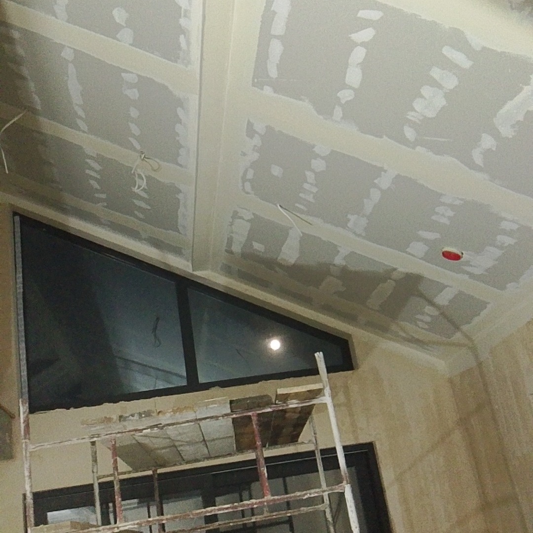 Sufit z płyt karton-gips, częściowo wyszpachlowany, widoczne łączenia. Okno trójkątne w czarnej ramie. Fragment rusztowania. Prace wykończeniowe wnętrza.