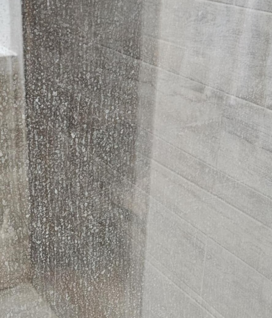 Porównanie: brudna, matowa szyba kabiny prysznicowej po lewej i czysta, błyszcząca po prawej, z widoczną strukturą płytek w tle. Efekt po czyszczeniu.