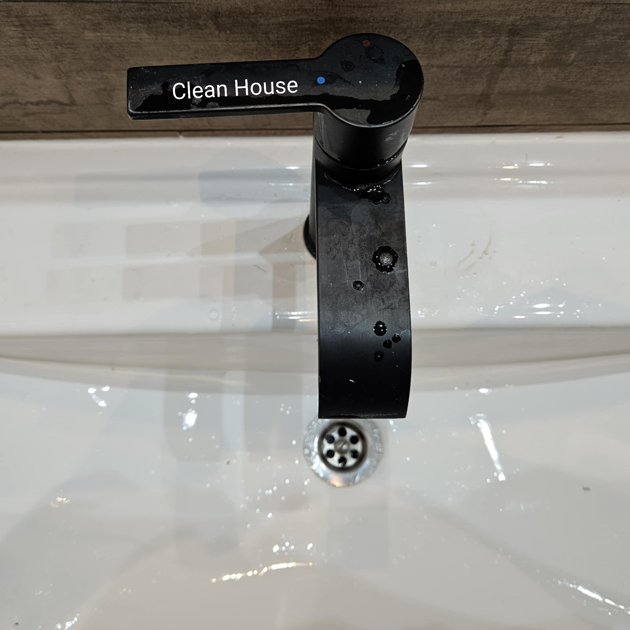 Błyszcząca, czarna bateria umywalkowa z kroplami wody na białej, czystej umywalce. Widoczne ślady po czyszczeniu. Tło z drewnianą teksturą.