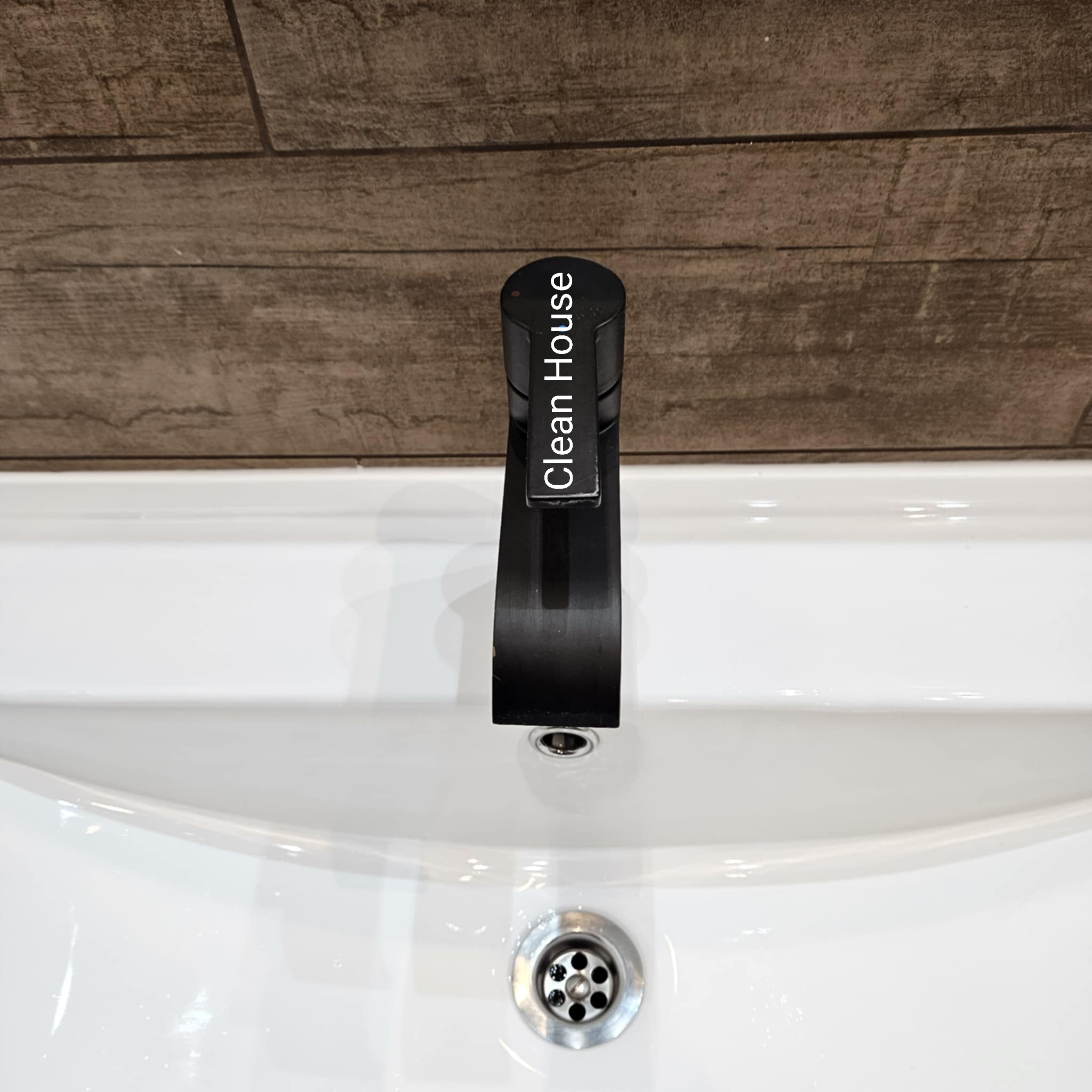Biała umywalka z czarną, minimalistyczną baterią z napisem 'Clean House' na tle drewnopodobnych płytek. Widok z góry, czysty i schludny.