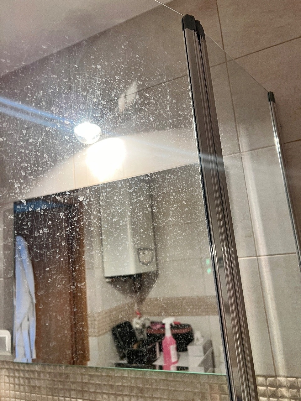 Zabrudzona kabina prysznicowa z widocznymi zaciekami i osadami, odbijająca fragment łazienki z bojlerem i akcesoriami. Potrzeba gruntownego czyszczenia.