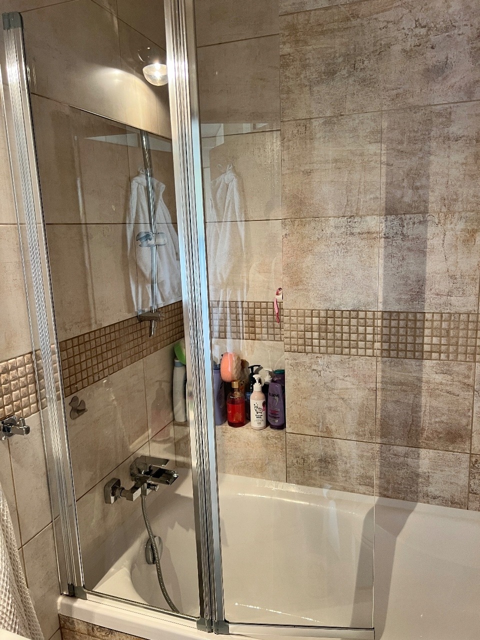 Czysta kabina prysznicowa z beżowymi płytkami i szklanymi drzwiami, widoczne kosmetyki i ręczniki. Bateria błyszczy, a na ścianie brak zacieków.