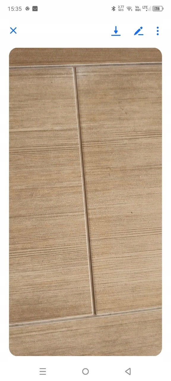 Beżowe płytki podłogowe z delikatną teksturą drewna i fugami. Widoczne drobne zabrudzenia na powierzchni płytek. Ujęcie z bliska, ukazujące detale wykończenia.