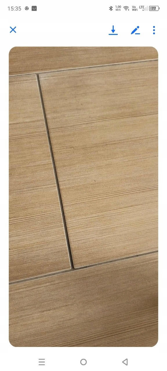 Beżowe płytki podłogowe z delikatnym wzorem drewna i fugami, ułożone w regularny wzór. Widoczne drobne zabrudzenia w fugach, co sugeruje potrzebę czyszczenia.