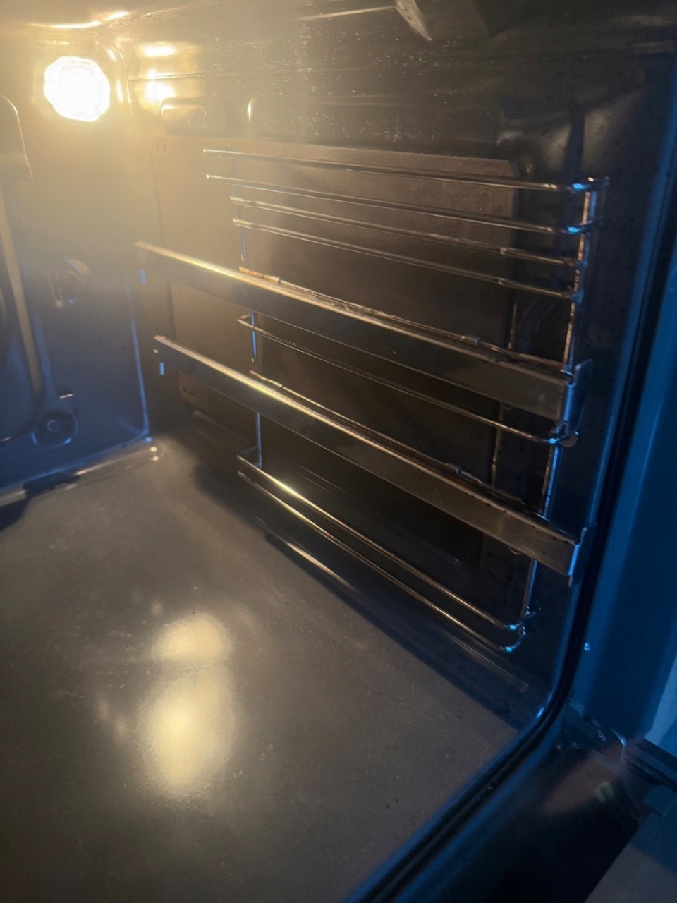 Wnętrze piekarnika z metalowymi półkami, oświetlone od góry. Widoczne ślady użytkowania na dnie i półkach. Piekarnik gotowy do czyszczenia.