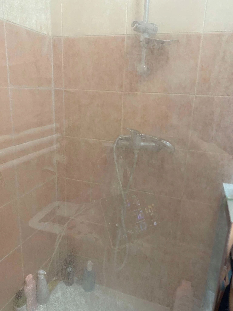 Zapuszczona kabina prysznicowa z osadem na szklanych drzwiach i płytkach w odcieniach beżu, widoczne butelki z kosmetykami. Wymaga gruntownego czyszczenia.
