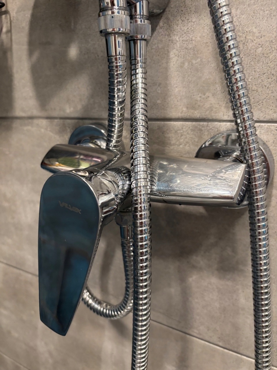Błyszcząca, chromowana bateria prysznicowa marki Vilmex na tle szarych płytek. Widoczne węże doprowadzające wodę. Ujęcie z bliska, podkreślające detale armatury.