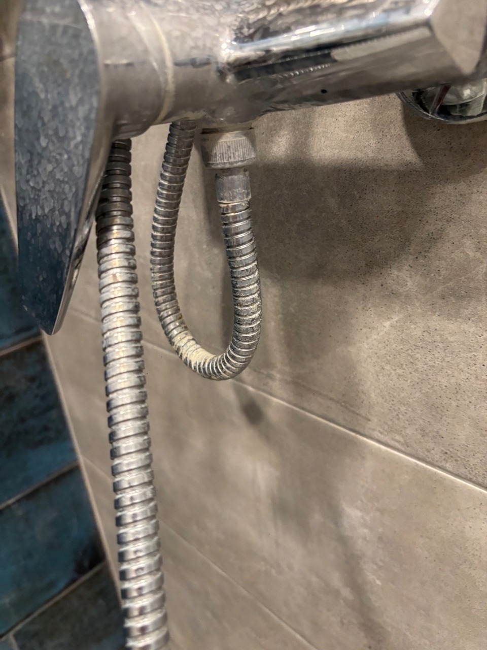 Zakamieniona bateria prysznicowa z widocznymi osadami na chromowanych elementach i wężach, na tle szarej, ceramicznej ściany w łazience.