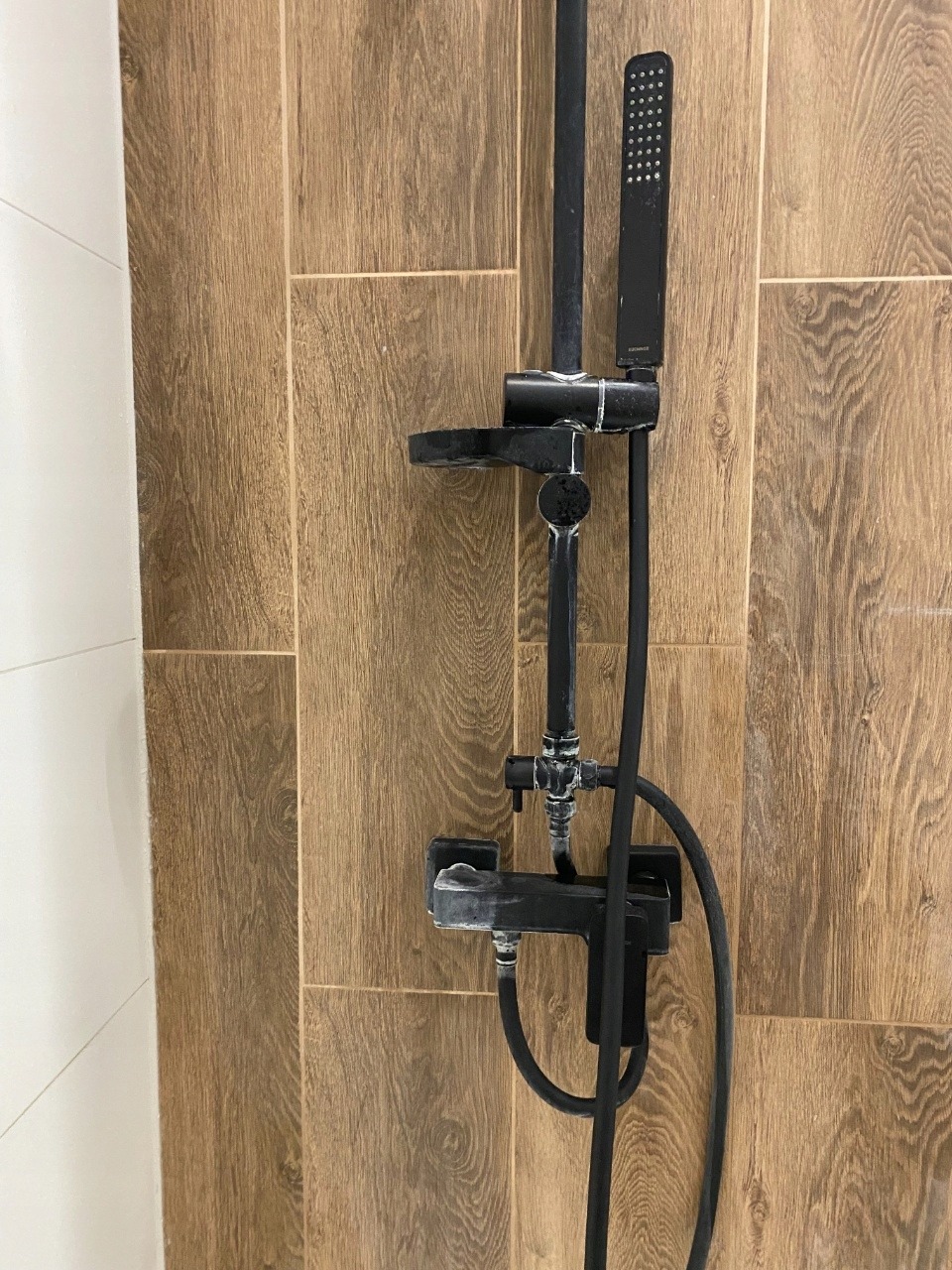 Czarny zestaw prysznicowy z osadami kamienia na tle drewnopodobnych płytek. Widoczne ślady po czyszczeniu, sugerujące potrzebę dokładniejszego sprzątania łazienki.