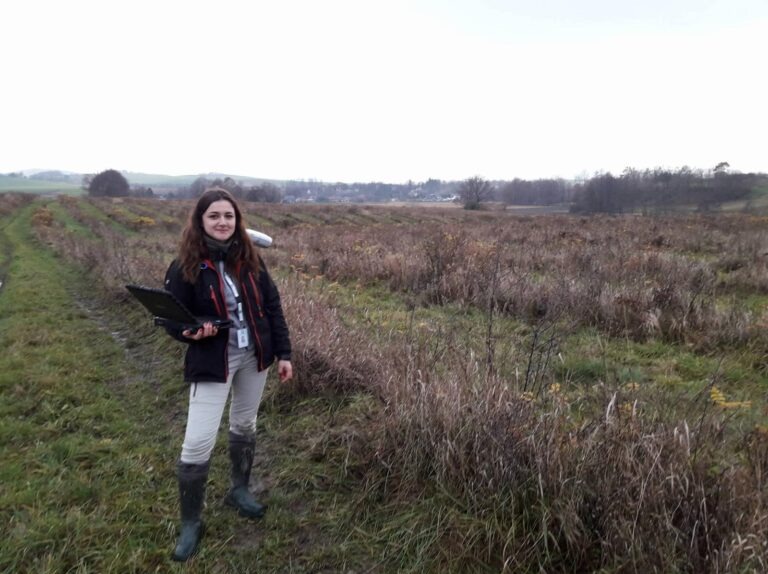 Kobieta geodeta z laptopem na tle rozległego terenu z niską roślinnością i odległym horyzontem, prawdopodobnie podczas pomiarów działki.