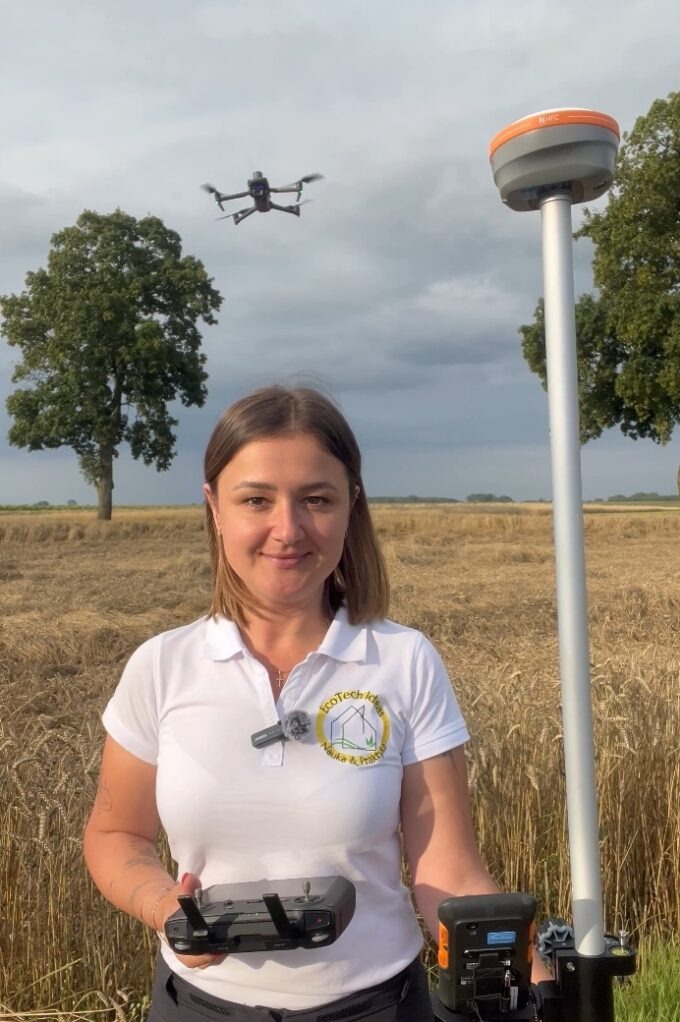 Geodeta z Poznania w terenie, trzyma kontroler drona. W tle lata dron i stoi odbiornik GNSS. Kobieta w koszulce polo z logo firmy.