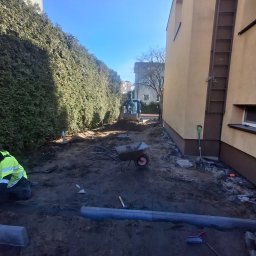 Artur Ulenberg - Przygotowanie terenu pod kostkę brukową: wykop, obrzeża, narzędzia, w tle koparka. Widok z perspektywy pracownika w odblaskowej kurtce, wzdłuż żywopłotu i budynku.