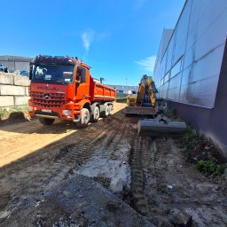 Artur Ulenberg - Pomarańczowa wywrotka Mercedes-Benz Arocs WMK Wandtke i żółta minikoparka na błotnistym placu budowy, przygotowujące teren przy ścianie hali przemysłowej.