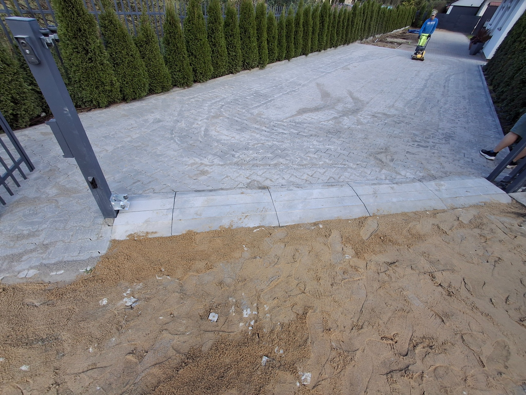 Ułożona szara kostka brukowa na podjeździe, widoczna brama wjazdowa i rząd tuj. W tle pracownik zagęszcza teren. Piasek czeka na rozprowadzenie.