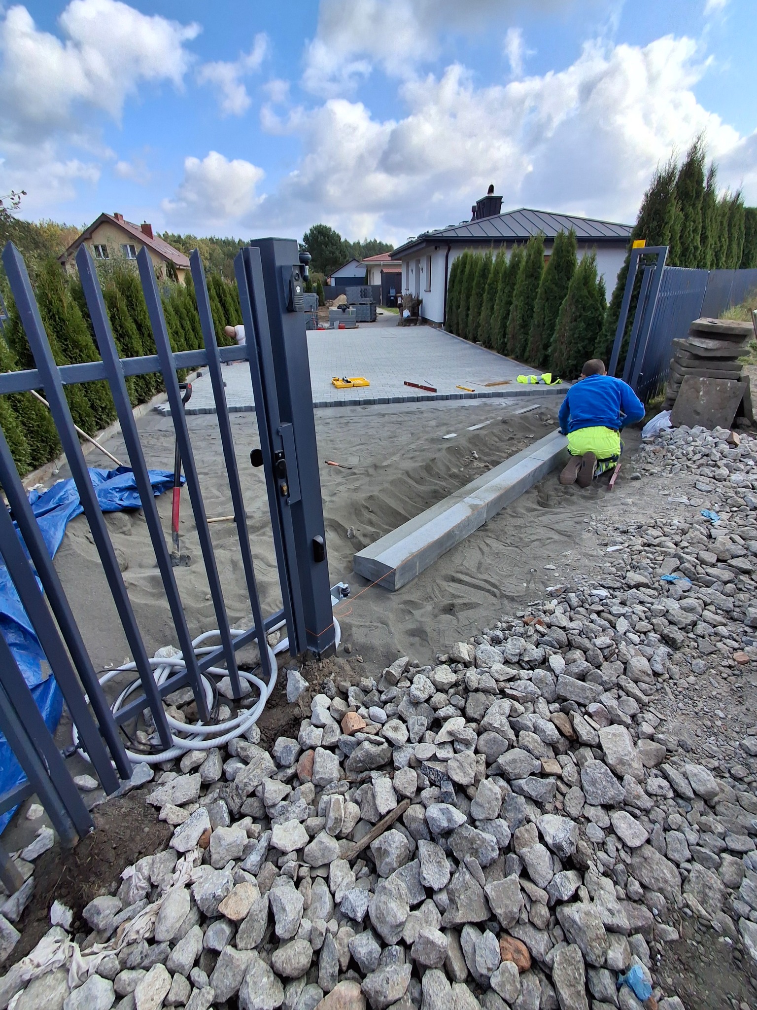 Montaż obrzeży betonowych przy posesji z szarą bramą. W tle ułożona kostka brukowa na podjeździe i dom z ciemnym dachem. Praca w toku, widoczne narzędzia i materiały.