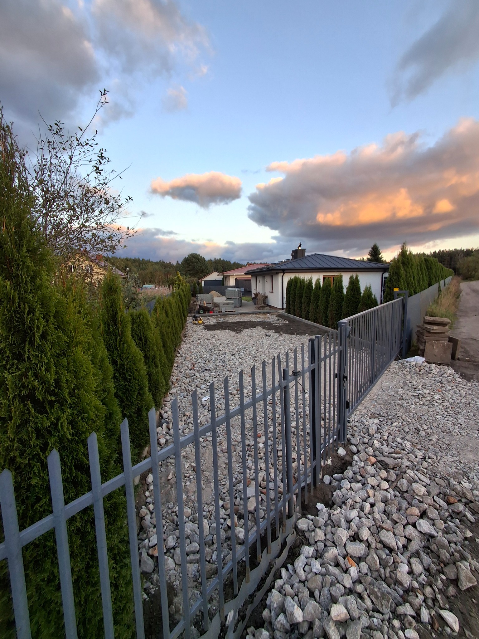 Montaż ogrodzenia panelowego na posesji z wysypanym kamieniem, widok na dom i tuje. Etap prac przygotowawczych, wieczorne światło.