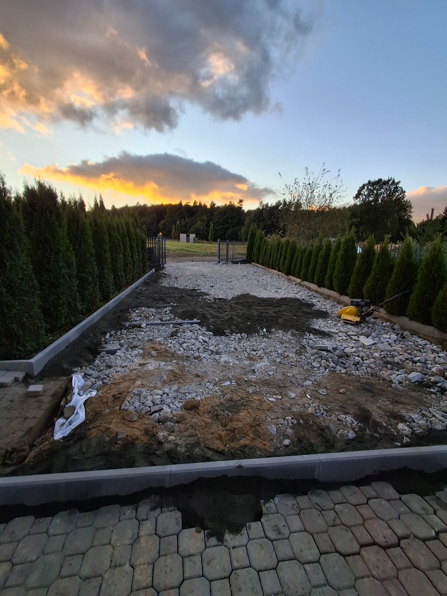 Etap przygotowania podłoża pod kostkę brukową: podsypka z kamienia i piasku, obrzeża betonowe, zagęszczarka w tle, rząd tuj wzdłuż ogrodzenia, zachodzące słońce.