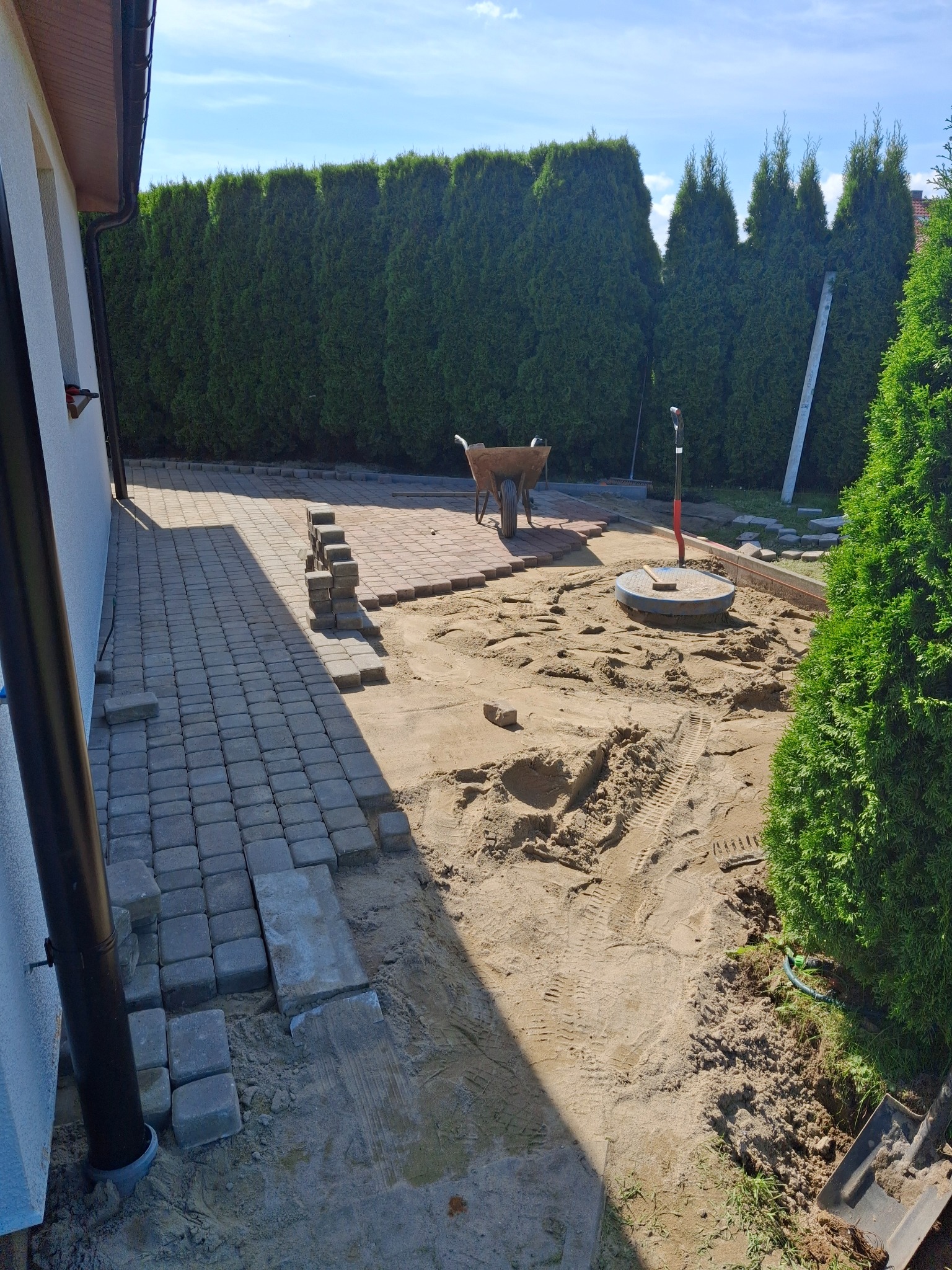 Fragment układanej kostki brukowej w ogrodzie, widoczna taczka i narzędzia, tło z tujami, częściowo ułożona nawierzchnia z szarej kostki.