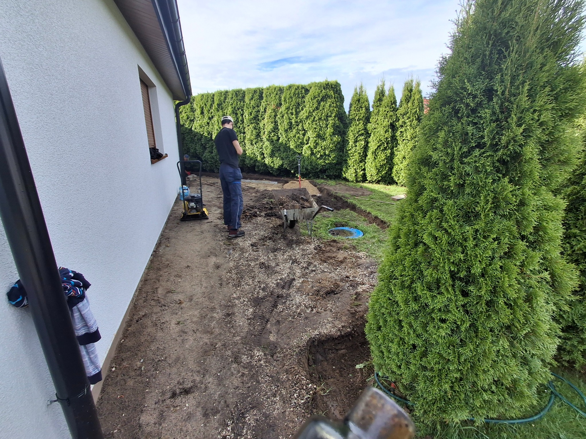 Przygotowanie terenu pod ścieżkę w ogrodzie: pracownik z ubijarką, wykopany rów, widoczne tuje i biała ściana budynku. Prace ziemne w toku.
