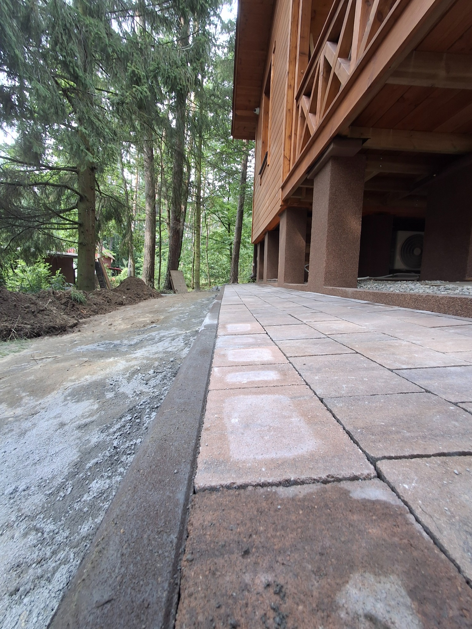 Ułożona kostka brukowa wzdłuż drewnianego domu na palach, widok z dołu. Częściowo przygotowany teren pod dalsze prace brukarskie. Otoczenie leśne.