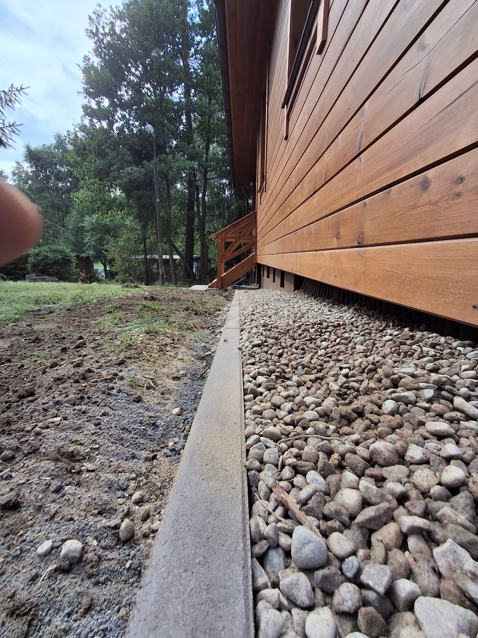 Wykończona podmurówka drewnianego domu z kamieniami i betonową obwódką, widok z poziomu gruntu, zieleń w tle.